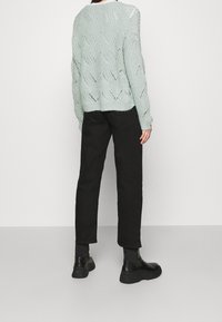 Pull en tricot vert clair avec un motif en vagues, porté avec un pantalon noir fuselé et des bottines noires, vu de dos.