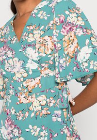 Vestido cruzado floral en tela azul verdoso. Presenta patrones vibrantes de flores en rosa, blanco y naranja. Destacan las mangas con volantes y el detalle de la cinta en la cintura.