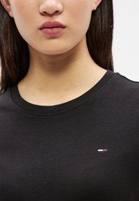 Kvinde iført en sort rundhalset trøje med et lille rødt, hvidt og blåt rektangulært logo på venstre bryst.