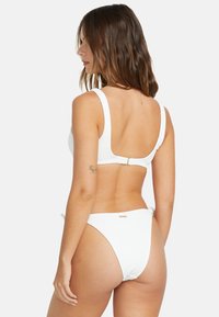 Set bikini bianco con tessuto testurizzato e spalline larghe. Il top ha una chiusura a gancio e i slip sono a vita bassa con un accento dorato.