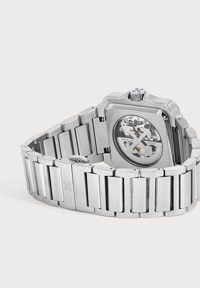 Montre-bracelet en acier inoxydable avec mouvement mécanique visible et bracelet à maillons métalliques, montrant le dos de la montre et la boucle sur fond blanc.