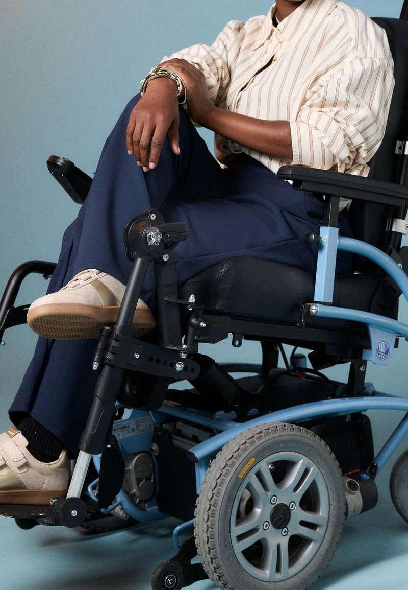 Personne aux jambes croisées, portant un pantalon bleu marine et une chemise rayée, assise dans un fauteuil roulant électrique bleu sur un fond neutre.