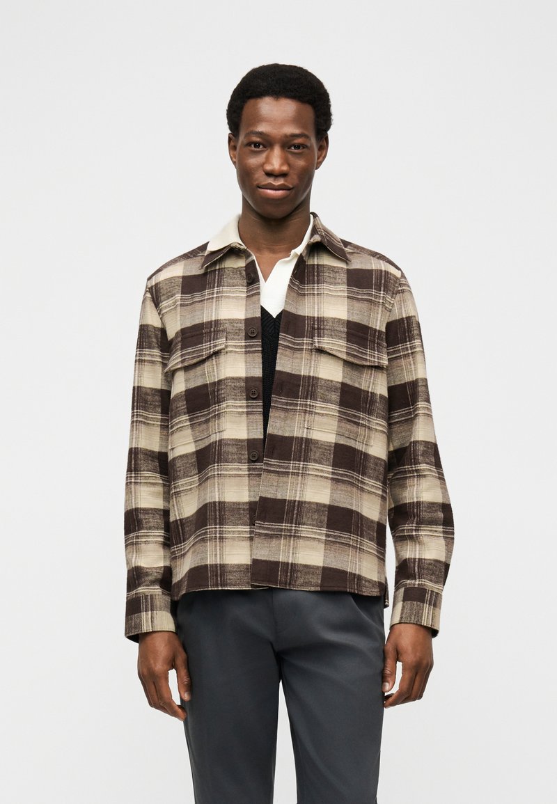 Junger Mann, der ein braun-beiges kariertes Hemd über einem schwarzen Pullover und einem weißen Hemd mit Kragen trägt, steht vor einem schlichten Hintergrund.