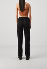 Kvinna som står med ryggen mot oss, iförd svarta lösa byxor, svarta klumpiga skor och en orange mönstrad crop top med tunna axelband.
