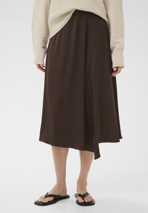 Sieviete tērpusies bēša džemperī, brūnā midi garuma svārkos ar asimetrisku apmali un melnās plakanās sandales ar starpupes siksnu uz balta fona.