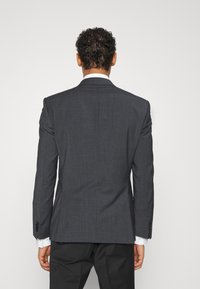 Mörkgrå kostymjacka med texturerad finish, strukturerade axlar, enkel slits och fyra-knappsmanschetter. Vit skjorta synlig under.