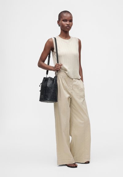 Femme portant un haut beige sans manches et un pantalon à jambes larges, tenant un sac à bandoulière en cuir noir, debout devant un fond uni.