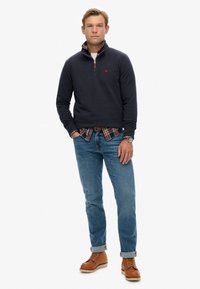 Marine pullover met halve rits, gecombineerd met een kraag van een geruite overhemd. Draagt bij lichte spijkerbroeken en bruine veterschoenen.