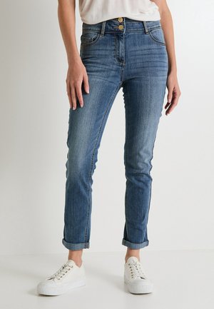 Kobieta w niebieskich, obcisłych dżinsach z wysokim stanem i podwijanymi nogawkami z podwójnym zapięciem na guziki oraz w białych sneakersach na platformie, stojąca na jednolitym tle.