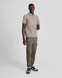 Camisa polo beige con tres botones y un logotipo amarillo. Pantalones cargo verde oliva con varios bolsillos. Zapatillas con un patrón marrón y crema.