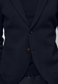 Närbild av en person som bär en mörk kavaj med två knappar knäppta över en mörk topp och byxor.