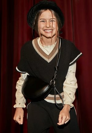 Gilet lavorato a maglia nero con scollatura a V sopra una camicetta crema. Borsa a spalla nera, cappello nero e pantaloni neri. Lo sfondo presenta tende rosse.