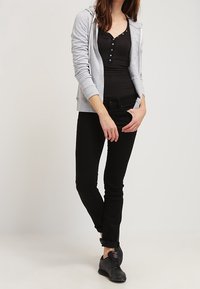 Sudadera gris con cremallera sobre una camiseta henley negra, combinada con jeans skinny negros y zapatos negros, creando un look casual en capas.