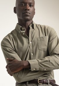 Camisa abotonada color verde oliva con un bolsillo en el pecho, fabricada en tela suave; cuenta con puños abotonados y un corte relajado. Cinturón trenzado marrón visible.