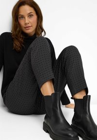 Zwarte leren enkellaarsjes met een dikke zool, gecombineerd met donkergekleurde broek met een geometrisch patroon. Model zittend tegen een witte muur.