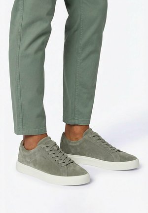 Persoon draagt matte groene suède sneakers met witte zolen en bijpassende groene broek, staand op een witte ondergrond.