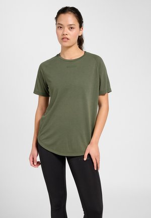 Junge Frau mit dunklen Haaren im Pferdeschwanz, die ein olivgrünes T-Shirt und schwarze Leggings trägt, steht vor einem schlichten hellen Hintergrund.
