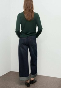 Jean large bleu foncé avec un revers gris, associé à un pull ajusté vert à manches longues, vu de dos. Chaussures noires.