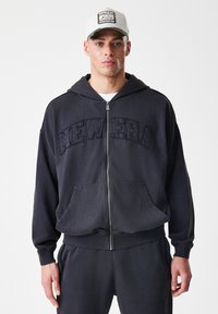 Felpa nera con zip e tasche frontali, caratterizzata da ricamo testurizzato "NEW ERA". Indossata con pantaloni della tuta grigi abbinati e un cappellino.