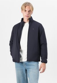 Marineblå zip-up jakke med oppstående krage, laget av lett materiale. Har en subtil logo og elastiske mansjetter. Kombinert med lyse jeans.