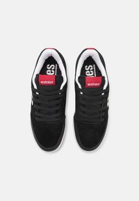 Black suede skate shoes with white accents, featuring a padded collar, black laces, and a red logo on the tongue and side. Rubber sole.