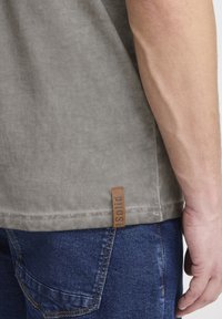 Grå bomulls t-skjorte med korte ermer, med en teksturert finish og et brunt lærmerke nederst. Brukt sammen med denim jeans.