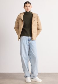 Krémová prošívaná bunda s kapucí, zelený rolák, světle modré oversized džíny a bílé tenisky. Jednoduchý design s minimálními akcenty.