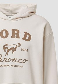 Beżowy kaptur z przytartym brązowym graficznym wzorem przedstawiającym bronco oraz napisem: "FORD Bronco 1966 Dearborn, MICHIGAN." Miękka tkanina.