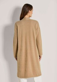 Eine Frau mit langen braunen Haaren, die einen langen, camel-farbenen, flauschigen Mantel trägt, steht vor einem einfarbigen hellen Hintergrund und schaut weg.