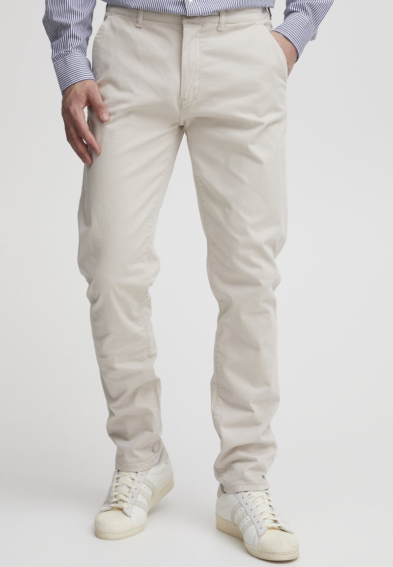 Beige Baumwollchinos mit schmalem Schnitt, frontalen Taschen und Reißverschluss, kombiniert mit weißen Sneakers mit drei schwarzen Streifen.
