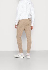 Pantalones de chándal beige con un patrón de logo de Nike en tonos repetidos, combinados con una sudadera blanca con capucha que presenta un diseño de logo similar.