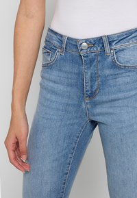 Jean en denim bleu clair avec une taille haute, cinq poches et des surpiqûres orange contrastantes. Le matériau semble doux avec une légère texture.