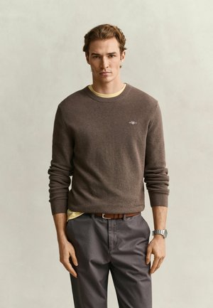 Junger Mann mit braunem Strickpullover über gelbem Hemd, grauer Hose, braunem Gürtel und silberner Uhr steht vor einem schlichten, hellen Hintergrund.