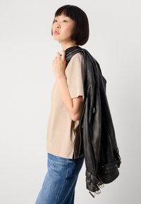 Donna con capelli corti e scuri che indossa una camicia beige e jeans blu, tiene una giacca di pelle nera sulla spalla e guarda verso l'alto.