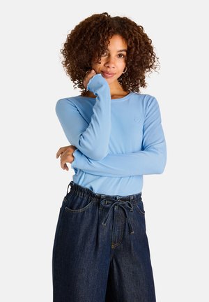 Frau mit lockigem Haar trägt ein hellblaues Langarmshirt und dunkle Jeans, posiert mit einer Hand im Gesicht und einem leichten Lächeln.