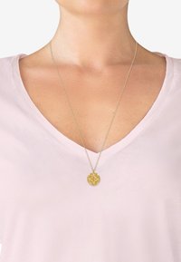 Guld vedhæng halskæde med indviklet design, hængende på en delikat kæde, båret over en lyserød v-hals skjorte.