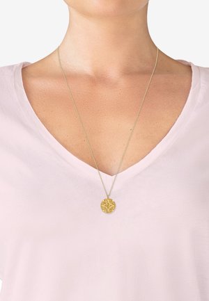 Goldener Anhänger mit kompliziertem Design, aufgehängt an einer feinen Kette, getragen über einem hellrosa V-Ausschnittshirt.