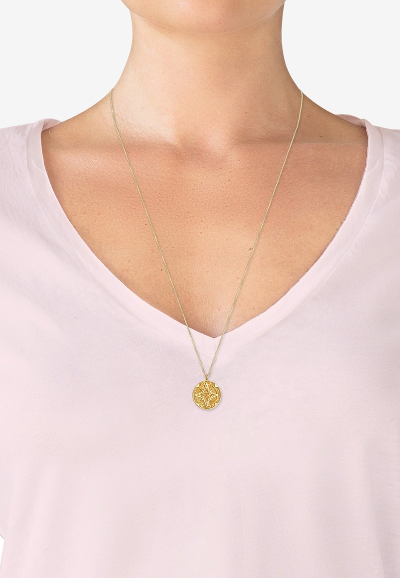 Collier en pendentif en or avec un design complexe, suspendu à une chaîne délicate, porté sur un t-shirt en v léger rose.