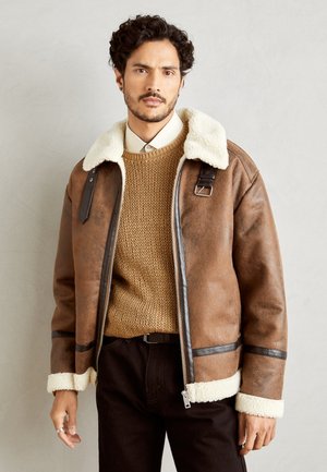 Veste en cuir synthétique marron avec un col et des poignets en shearling blanc, présentant des accents noirs et une fermeture éclair. Portée par-dessus un pull en tricot beige.