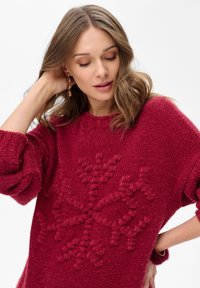 Czerwony sweter z dzianiny z fakturowanym wzorem płatka śniegu, szerokimi rękawami i zaokrąglonym dekoltem. Miękki, puszysty materiał o przytulnym wyglądzie.