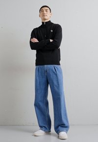 Fekete cipzáras pulóver, kicsi logóval a mellrészen, laza világoskék farmernadrággal és fehér tornacipővel párosítva.