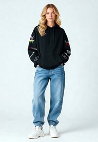 Sudadera negra con diseños bordados coloridos en las mangas, combinada con vaqueros anchos de color azul claro y zapatillas blancas. Tejido suave, ajuste casual.