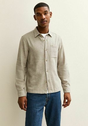 Camicia di lana beige con bottoni frontali, maniche lunghe, colletto e taschino sul petto. Indossata sopra una maglietta bianca e abbinata a jeans blu.