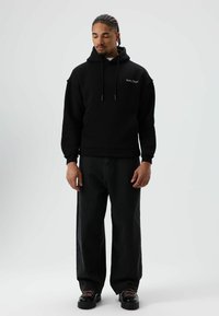 Zwarte hoodie met een voorzak en een capuchon met trekkoord, gecombineerd met losse zwarte jeans en stevige zwarte schoenen met gekleurde veters.