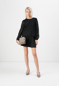 Black long-sleeved blouse with textured pattern, paired with a pleated black skirt. Holding a beige snake-print handbag and wearing grey heels.