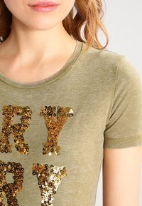T-shirt in cotone verde oliva con maniche corte, caratterizzato da grandi lettere in paillettes oro e marrone che formano la parola "VERY" sulla parte anteriore. Texture liscia.