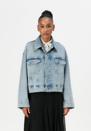 Lichtblauwe denimjas met een cropped ontwerp, grote zakken, knoopsluiting en brede mouwen. Gedragen over een zwarte rok en een wit overhemd met kraag.