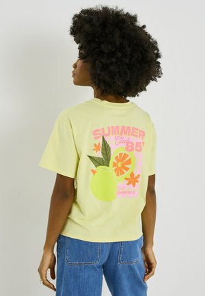 Mujer con cabello rizado que lleva una camiseta amarilla con frutas rosas y naranjas y el texto "SUMMER Club 85" en la espalda, combinada con vaqueros azules.