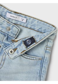 Jeans de mezclilla azul claro con herrajes metálicos, costuras en contraste y cierre de botón. La tela tiene una textura ligeramente desgastada.