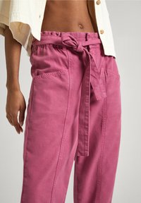 Persona con pantalones rosas de talle alto con cinturón atado, chaqueta beige corta y abdomen visible sobre un fondo neutro.